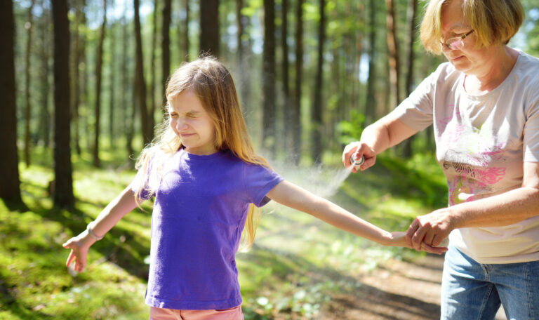 Middle age woman applying insect repellent to her granddaughter before forest hike beautiful summer day. Protecting children from biting insects at summer. Active leisure with kids.