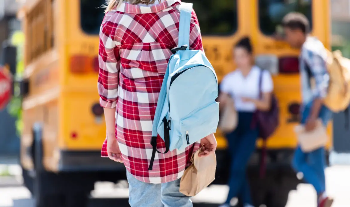 rear view of teen schoolgirl walking to classmates leaning on school bus