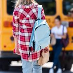 rear view of teen schoolgirl walking to classmates leaning on school bus