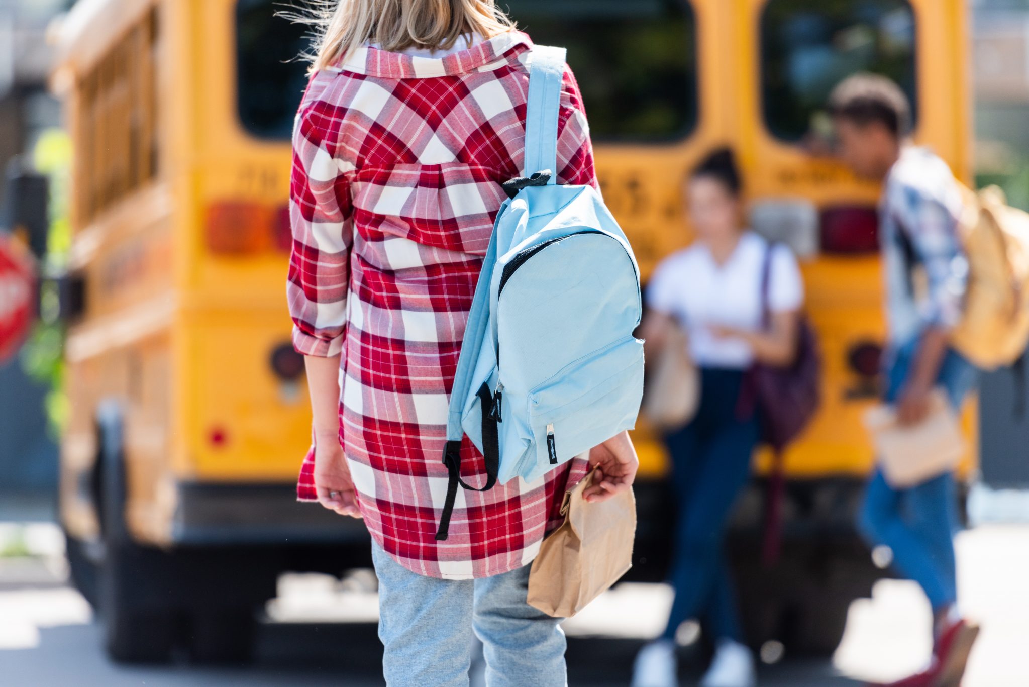 rear view of teen schoolgirl walking to classmates leaning on school bus