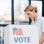 side view of attractive woman voting near stand with vote lettering and american flag