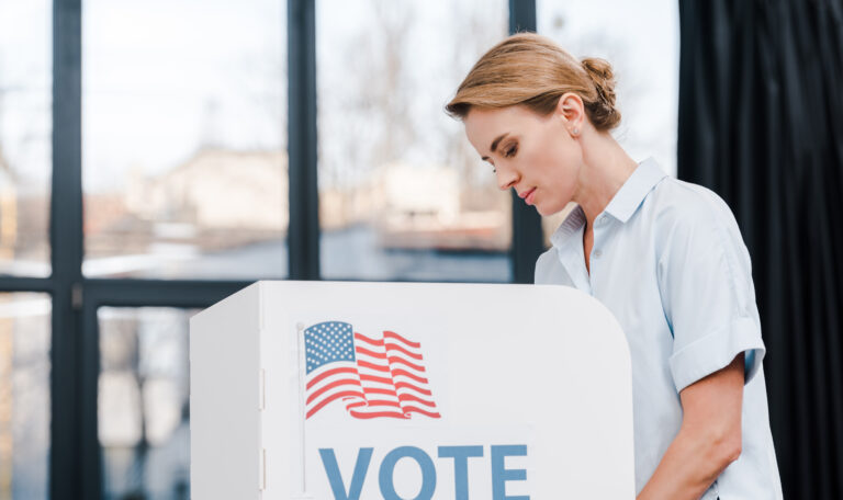 side view of attractive woman voting near stand with vote lettering and american flag