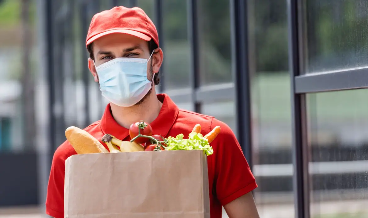 Courier in medical mask holding shopping bag with fresh vegetables on urban street