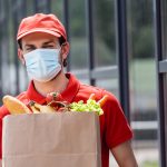 Courier in medical mask holding shopping bag with fresh vegetables on urban street
