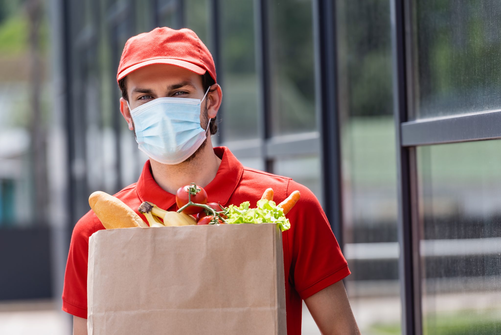 Courier in medical mask holding shopping bag with fresh vegetables on urban street