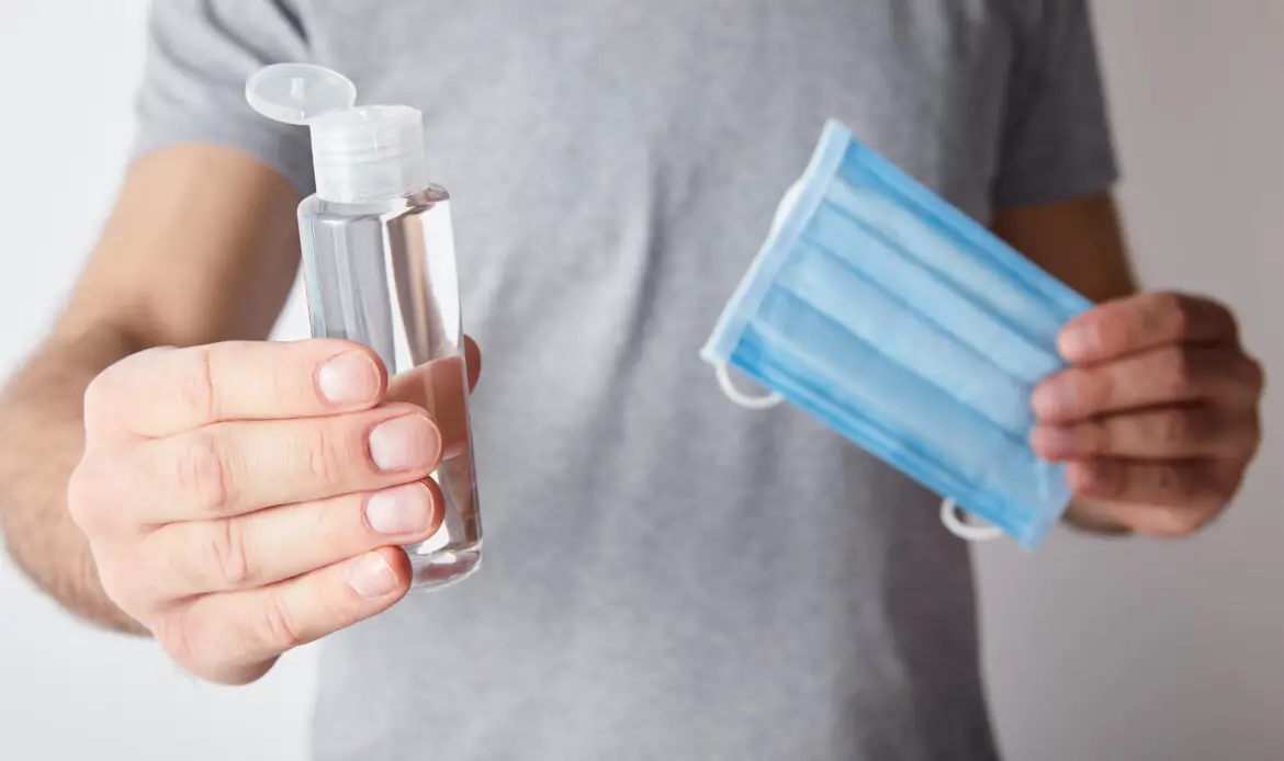 cropped view of man holding gel hand sanitizer in bottle and medical mask on grey background