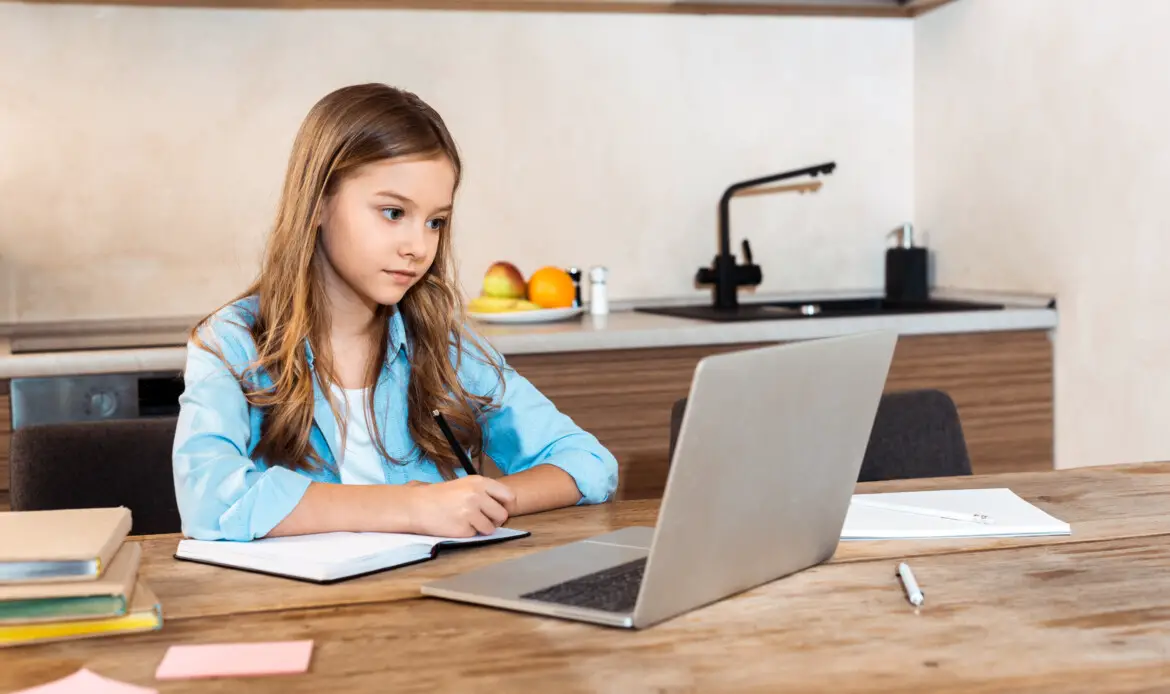 selective focus of cute kid writing in notebook near laptop while e-learning at home