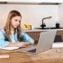 selective focus of cute kid writing in notebook near laptop while e-learning at home