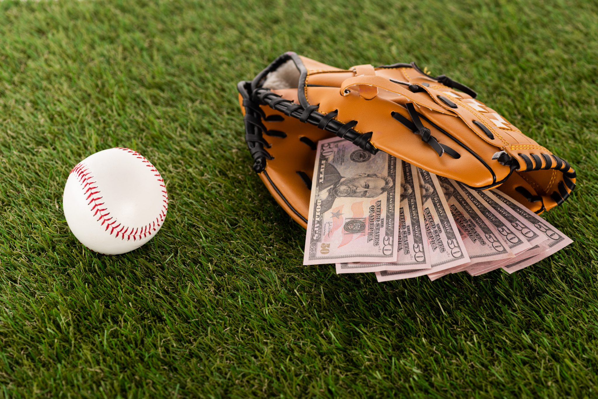 dollar banknotes in baseball glove near ball on green grass, sports betting concept