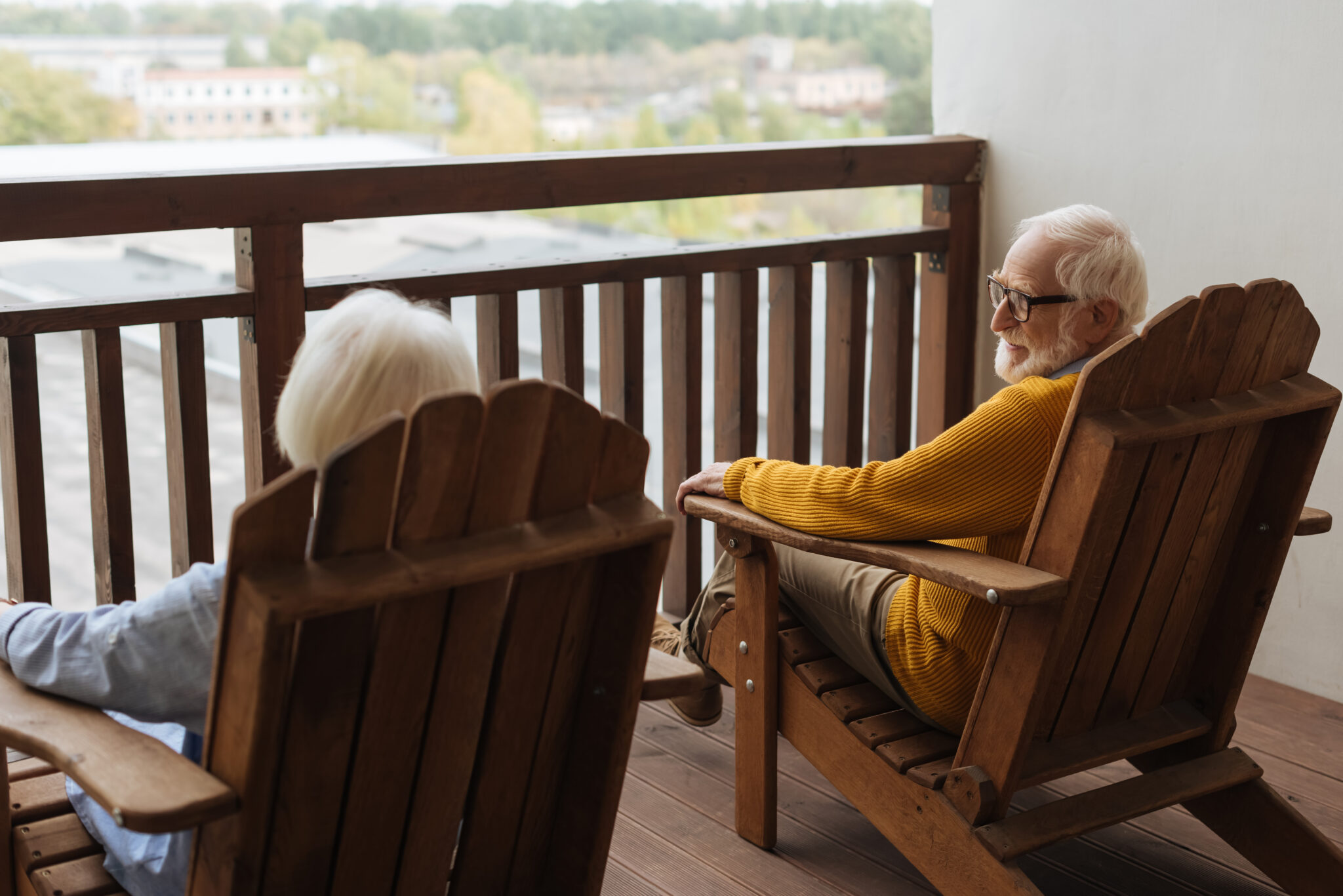 back view of wife with grey hair sitting near smiling husband in wooden armchair on blurred background on terrace