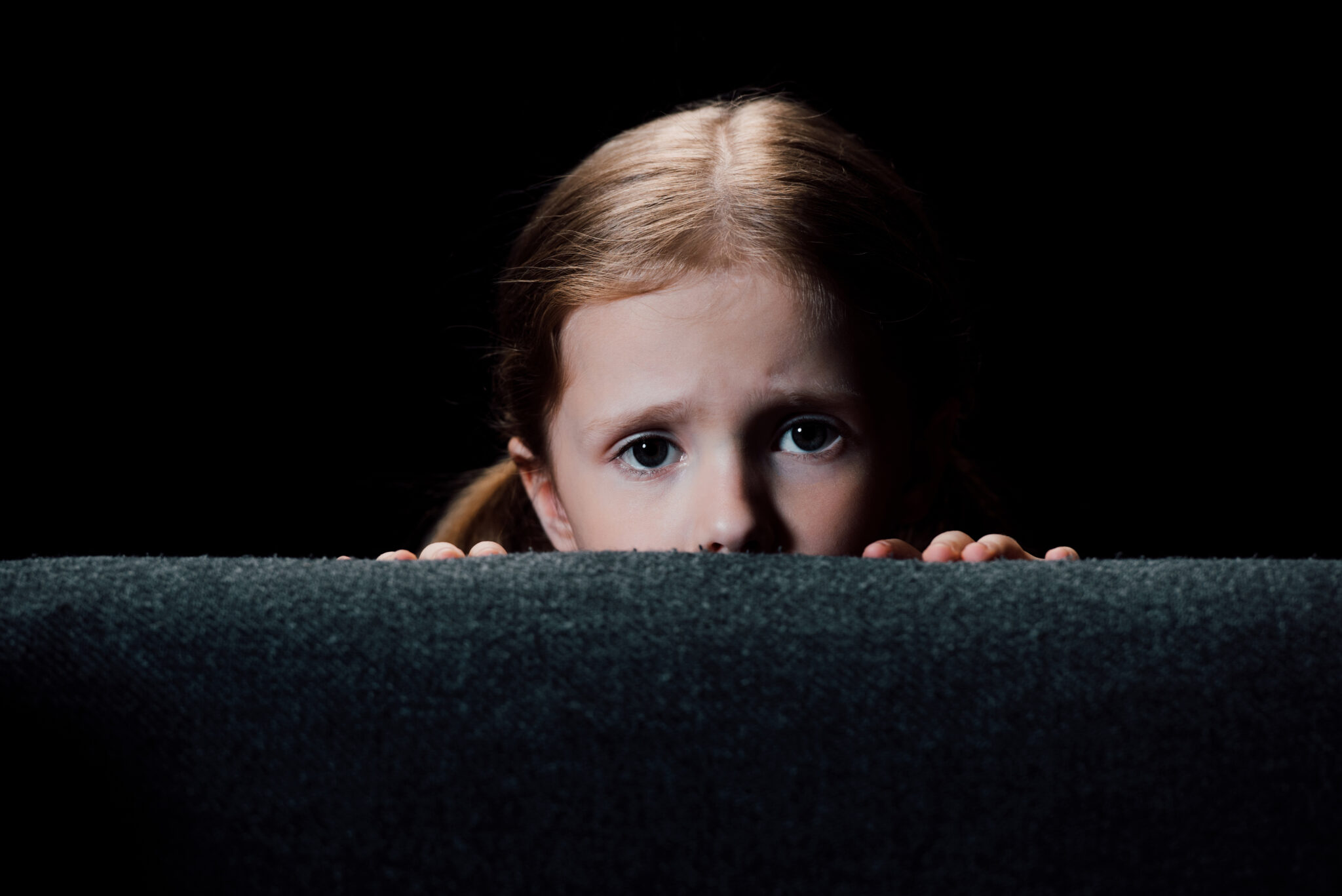 scared child hiding behind armchair and looking at camera isolated on black