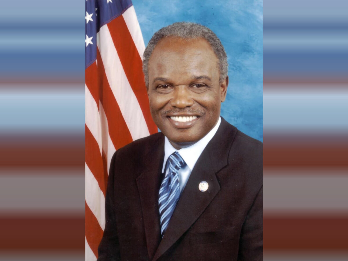 Portrait of a smiling man wearing a dark suit, light blue shirt, and blue striped tie, with an American flag in the background.