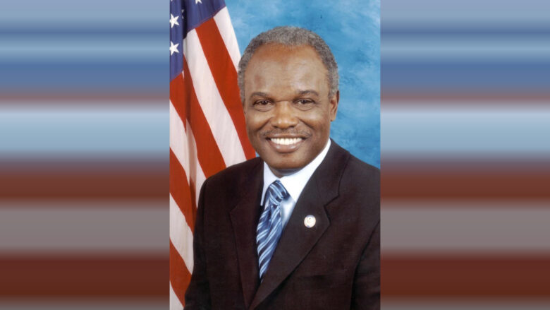 Portrait of a smiling man wearing a dark suit, light blue shirt, and blue striped tie, with an American flag in the background.