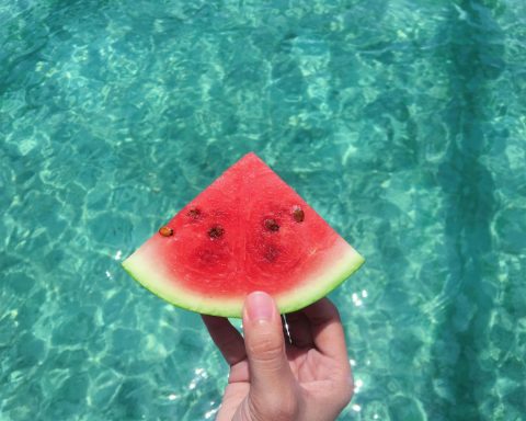 Hand Holding a Slice of Watermelon With Blue Swimming Pool Water in the Background