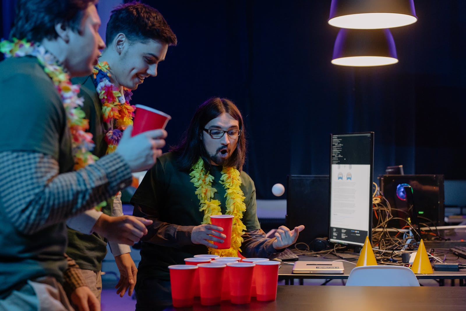 Three men play beer pong indoors, enjoying a lively gathering with red cups and leis.