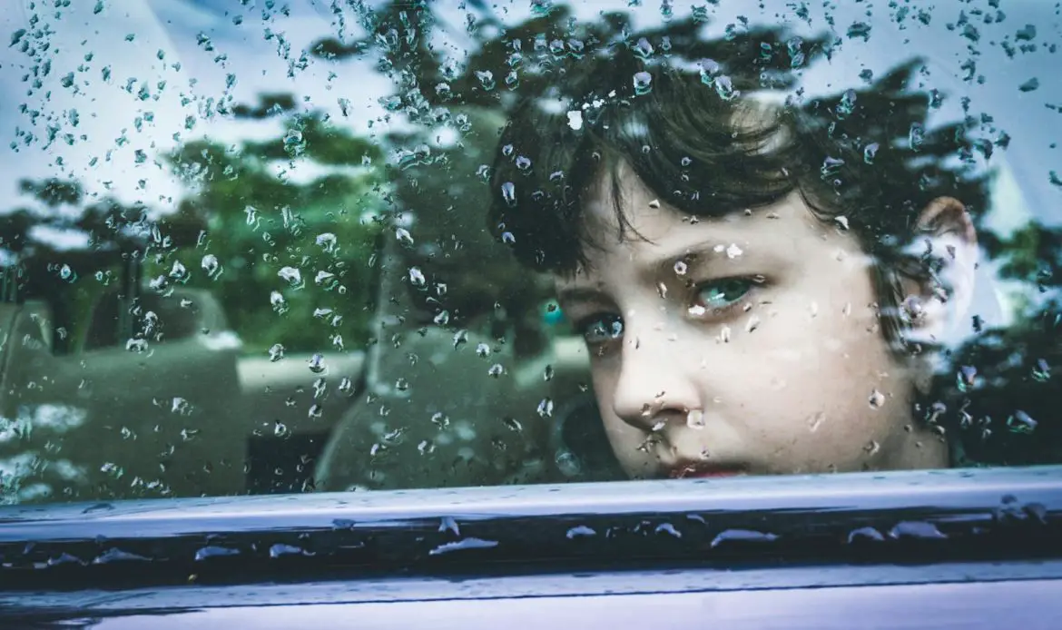 A child gazes contemplatively through a rain-marked car window, capturing a moment of introspection.