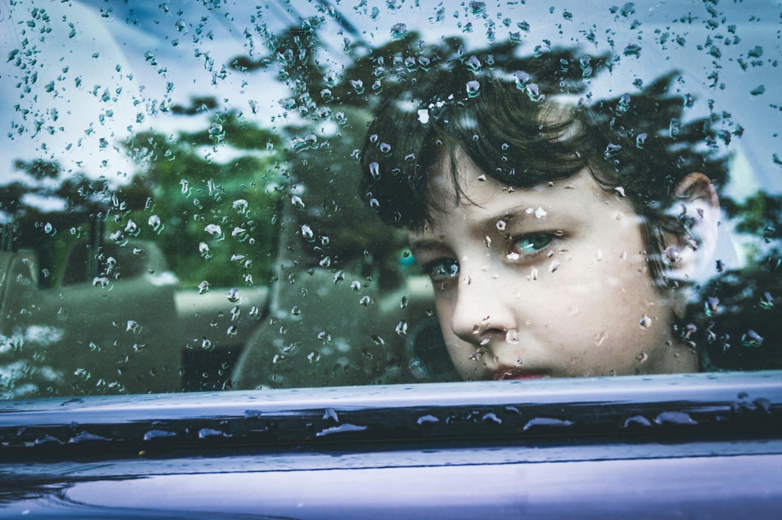 A child gazes contemplatively through a rain-marked car window, capturing a moment of introspection.