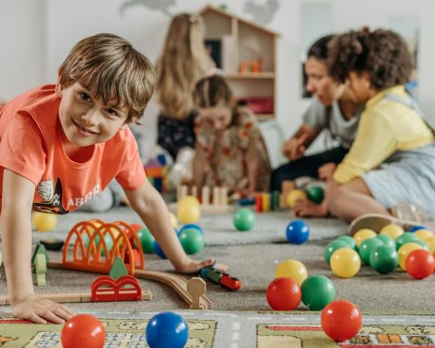 Happy children and a caregiver playing with colorful toys in a lively indoor playroom setting.