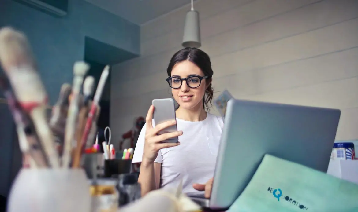 A woman using her phone at a desk, surrounded by art supplies and a laptop, in a creative workspace.