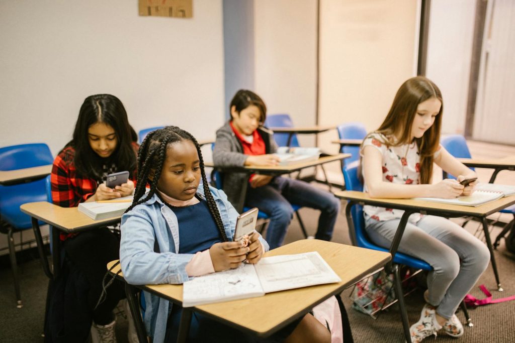 A group of four students in a classroom using smartphones and notebooks during a lesson.