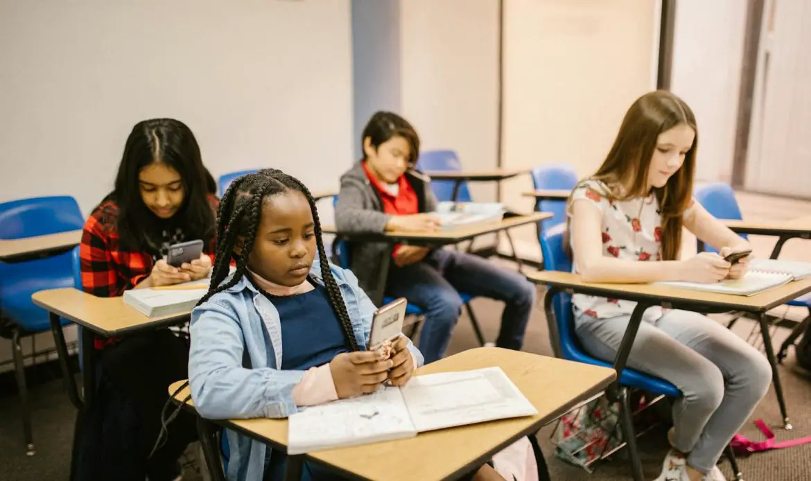 A group of four students in a classroom using smartphones and notebooks during a lesson.