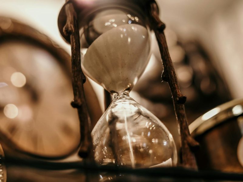 Close-up of a vintage hourglass with blurred antique clock background, evoking a timeless feel.