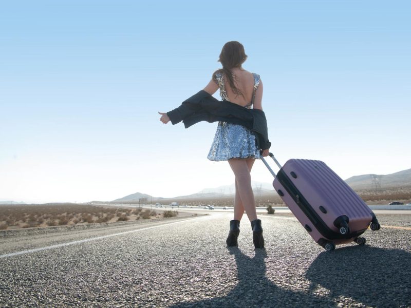 Woman in a sequined dress hitchhiking on a desert road with a pink suitcase. Fashion meets adventure.