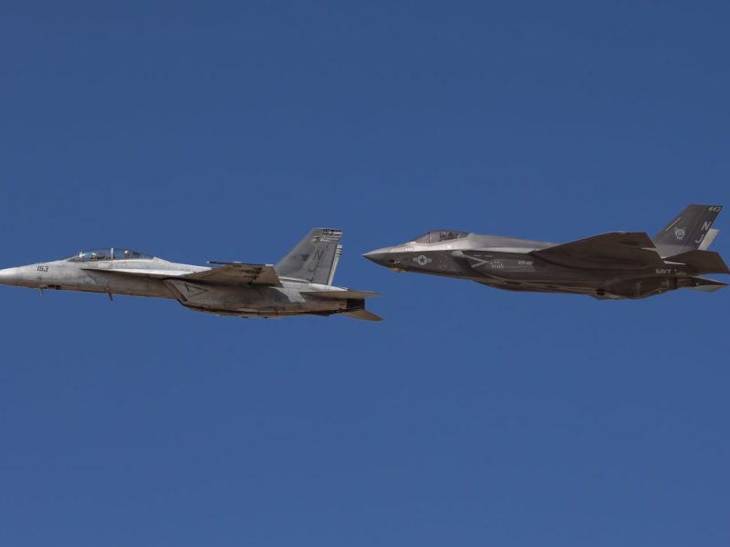 Two military jets, F-18 and F-35, flying in formation against clear blue sky.