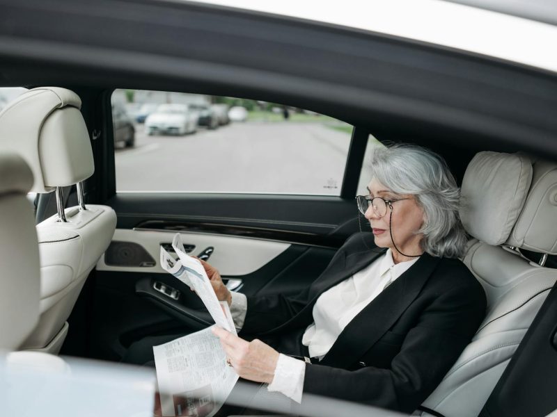 Elderly woman in a suit reading newspaper inside a luxury car.