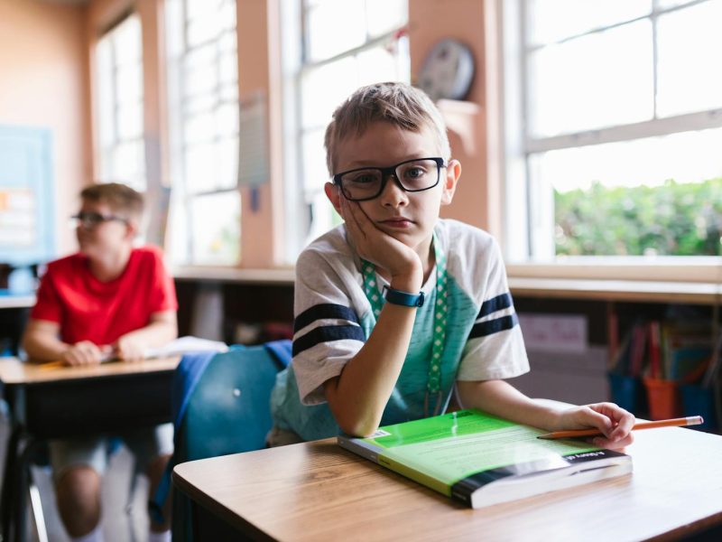 Young boy with glasses in a classroom, pen in hand, looking thoughtful.