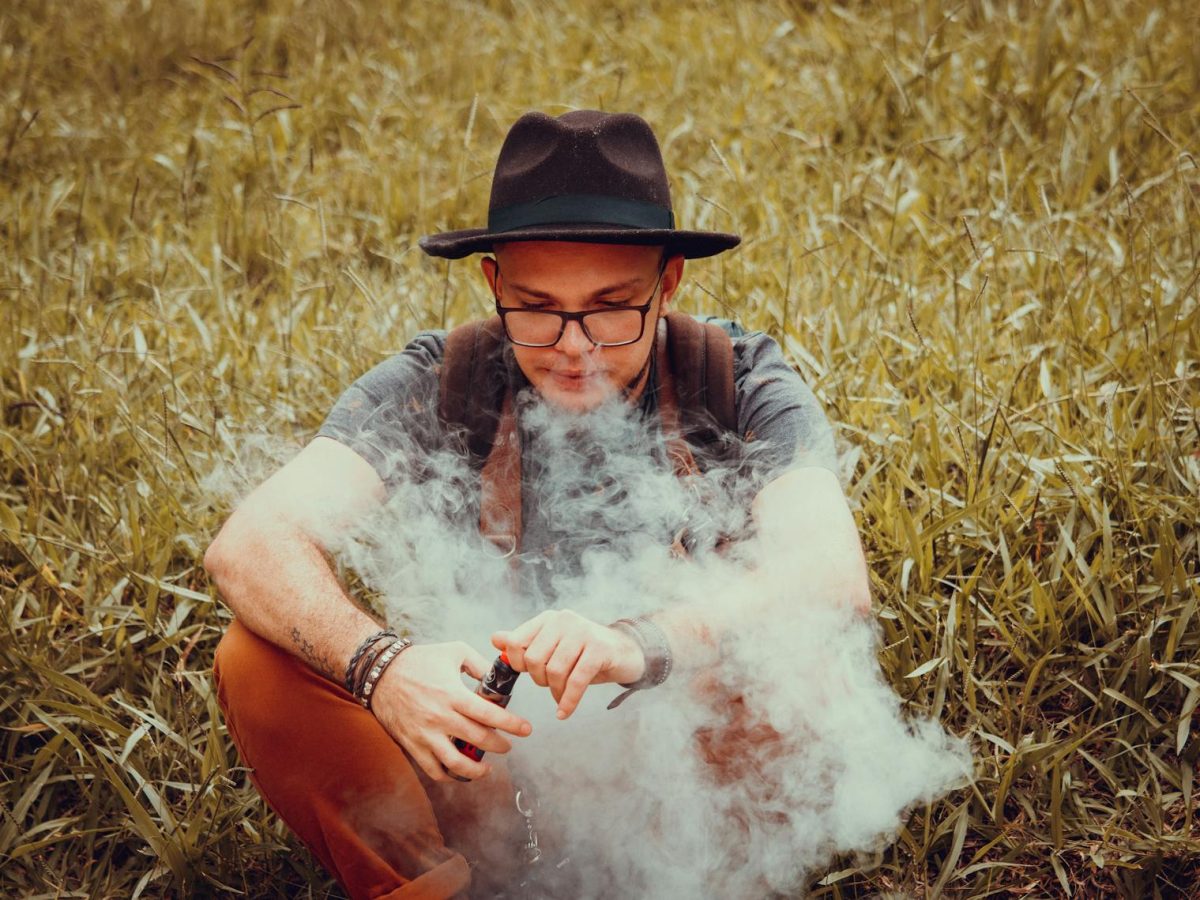 A man with a cowboy hat is sitting in a grass field, vaping on a clear day.