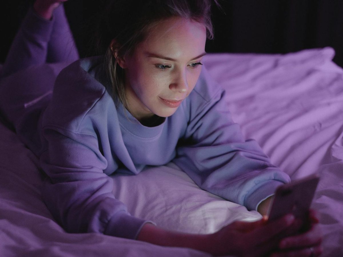 A woman in pajamas looking at her smartphone while lying on the bed under purple lighting, indoors at night.