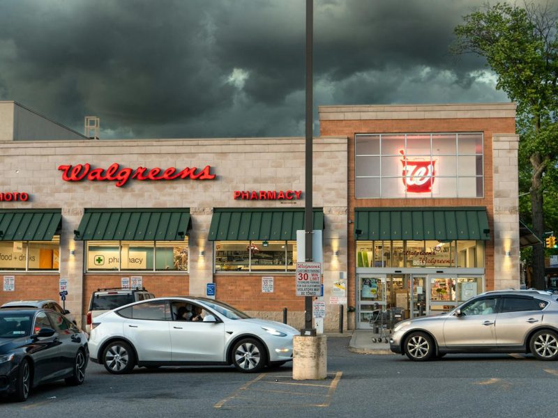 Exterior view of a Walgreens pharmacy under dark storm clouds with cars in the parking lot.