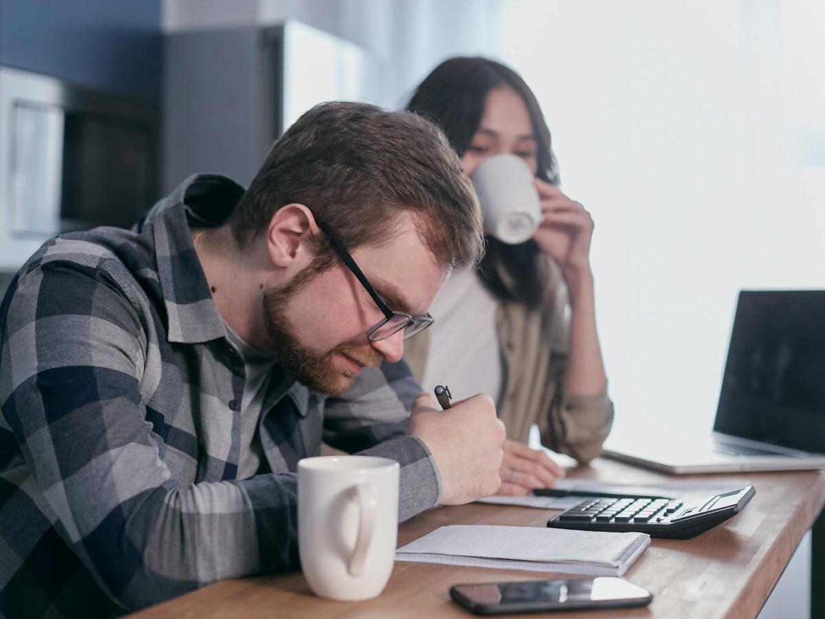 A couple sits at a kitchen table, reviewing finances with a laptop and calculator.