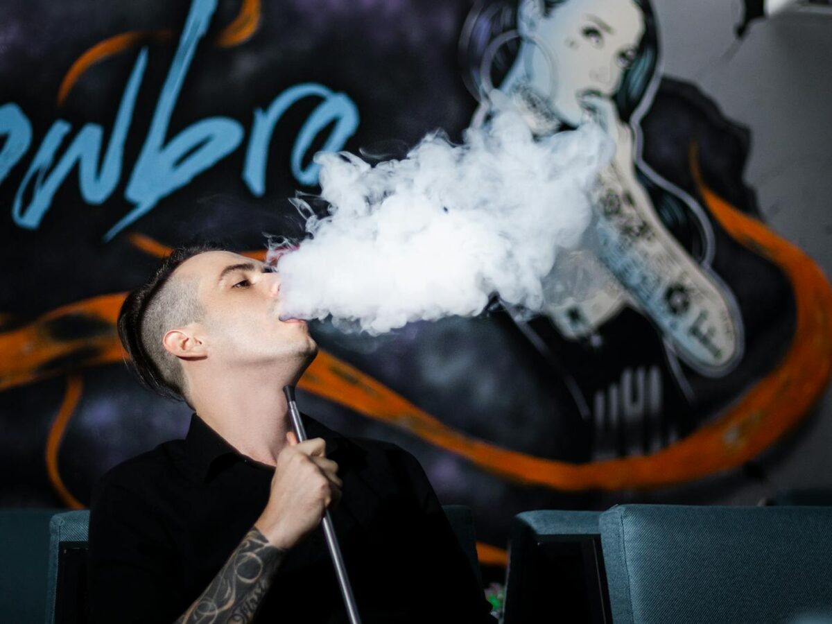 A man with tattoos vapes indoors, creating a smoke cloud against a mural backdrop.