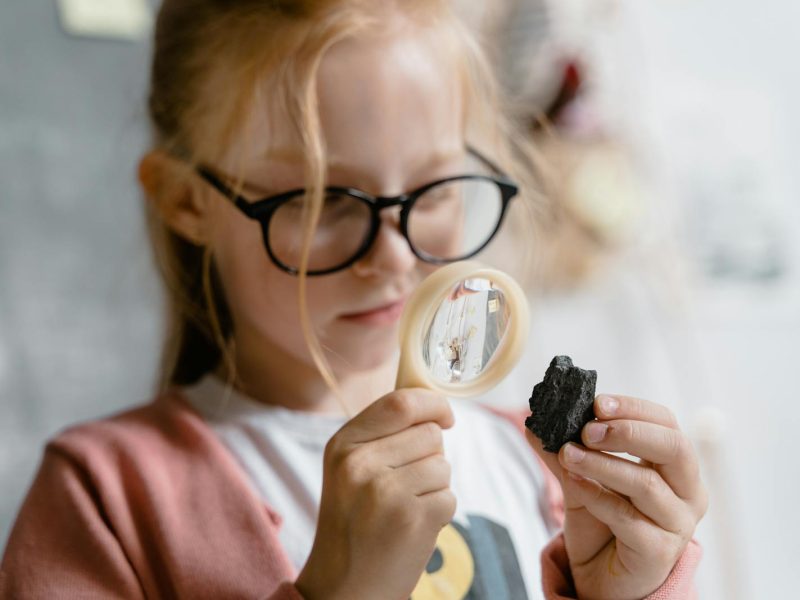 Young girl studies a rock closely with a magnifying glass, conveying curiosity and learning.