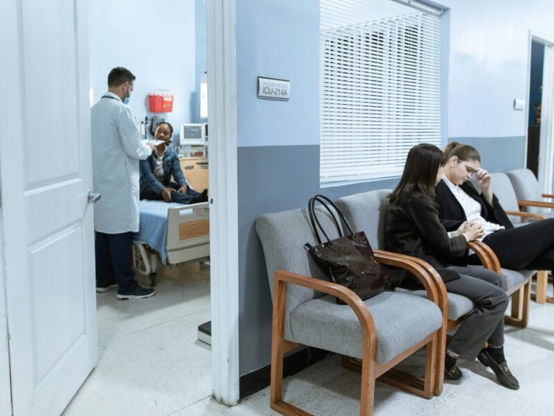 Doctor with patient in ICU, women in waiting area showing concern.
