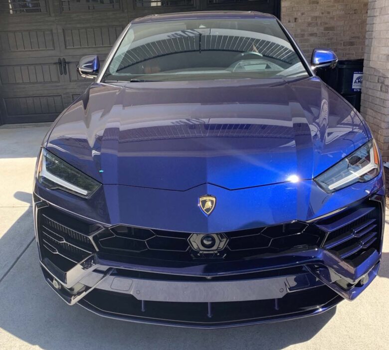 Front view of a blue Lamborghini Urus parked on a driveway in front of a black garage door. The car features sharp, angular headlights and a prominent Lamborghini emblem on the hood. The surface of the car is glossy, reflecting the surrounding environment.