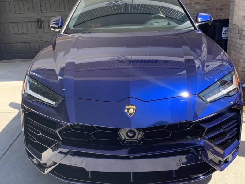 Front view of a blue Lamborghini Urus parked on a driveway in front of a black garage door. The car features sharp, angular headlights and a prominent Lamborghini emblem on the hood. The surface of the car is glossy, reflecting the surrounding environment.