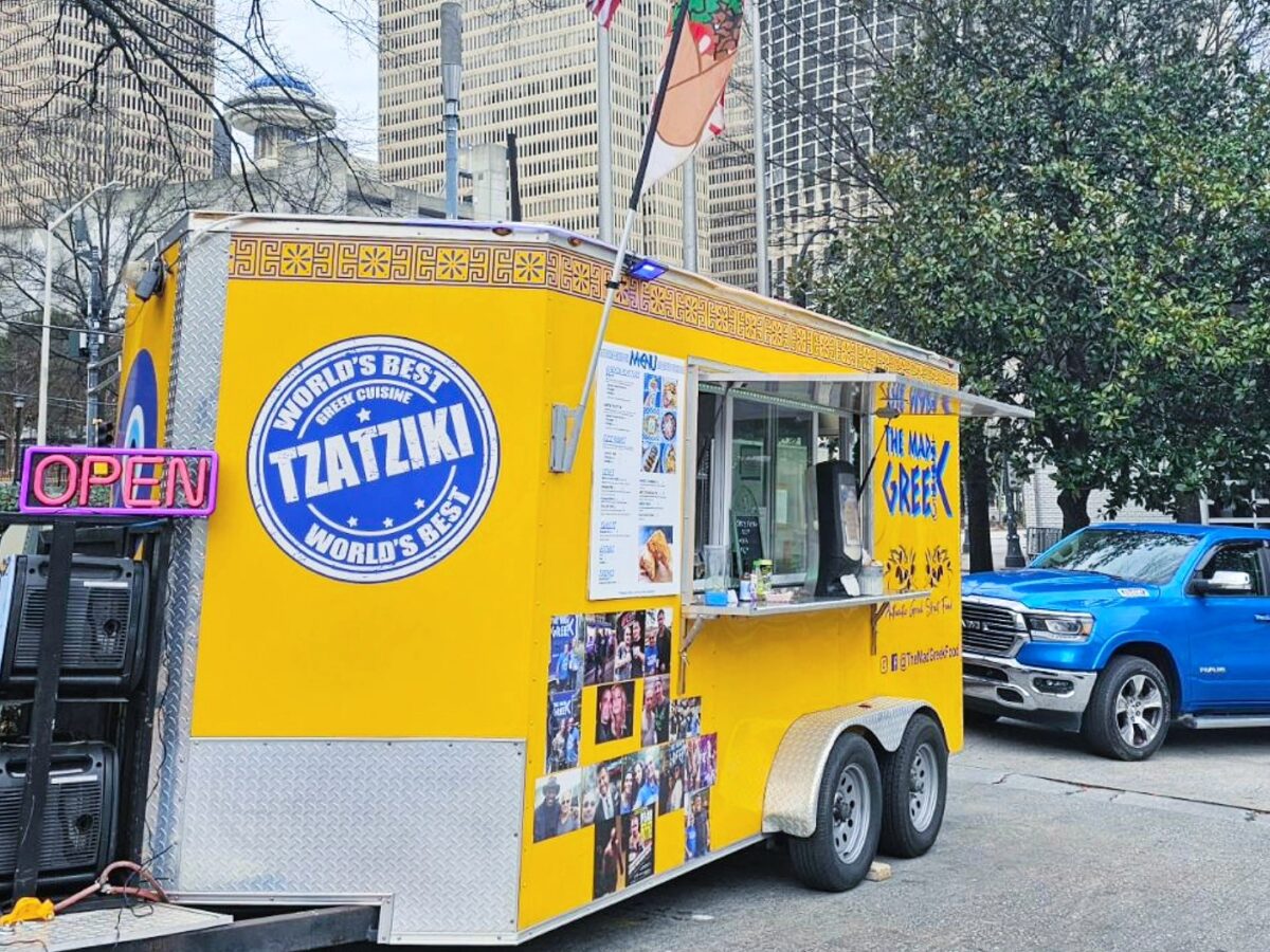 Yellow food truck with "World's Best Tzatziki" and "The Made Greek" branding, serving Greek cuisine, parked on a city street with tall buildings and trees in the background. A neon "OPEN" sign is visible on the side, along with a menu and photos of people. A blue pickup truck is parked nearby.
