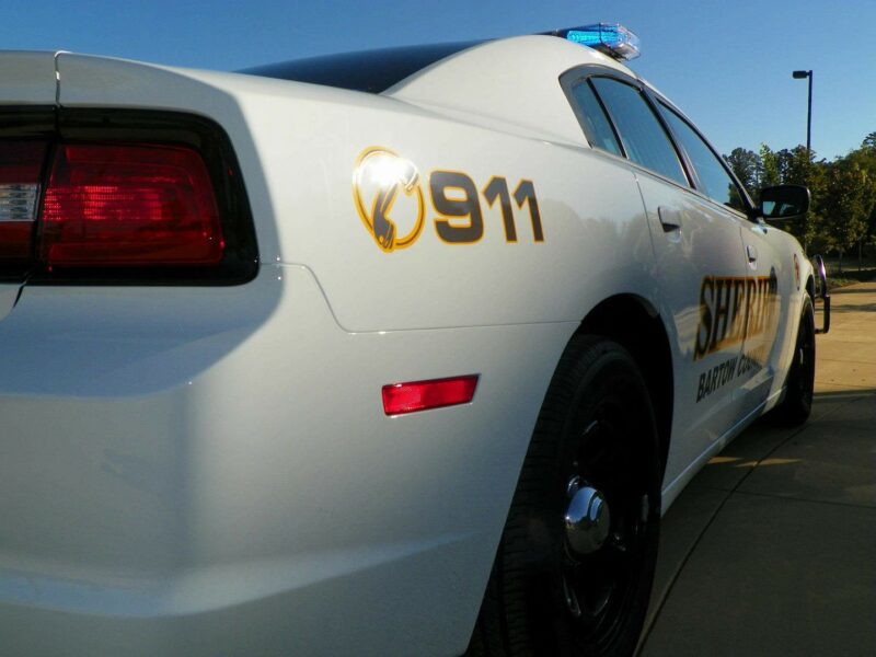 White Barton County sheriff's patrol car with "911" and sheriff insignia on the rear side, black wheels, and a blue emergency light on the roof, parked on a paved surface under a clear blue sky.