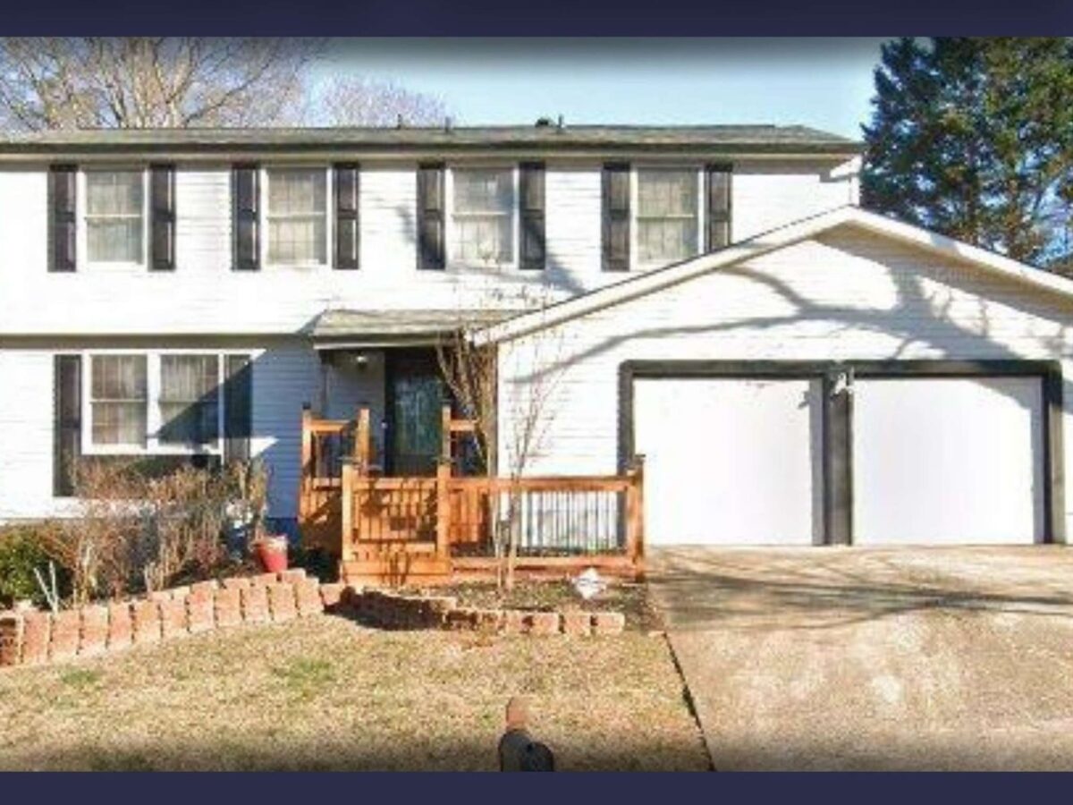 Two-story white house with black shutters and a double garage with white doors and black trim. The front yard has a small wooden porch with railings, a brick-bordered garden bed, and a concrete driveway. Trees and bushes surround the property. A Roswell, Georgia Police Department badge is visible in the bottom right corner.