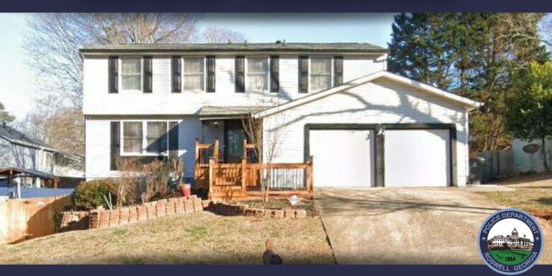 Two-story white house with black shutters and a double garage with white doors and black trim. The front yard has a small wooden porch with railings, a brick-bordered garden bed, and a concrete driveway. Trees and bushes surround the property. A Roswell, Georgia Police Department badge is visible in the bottom right corner.