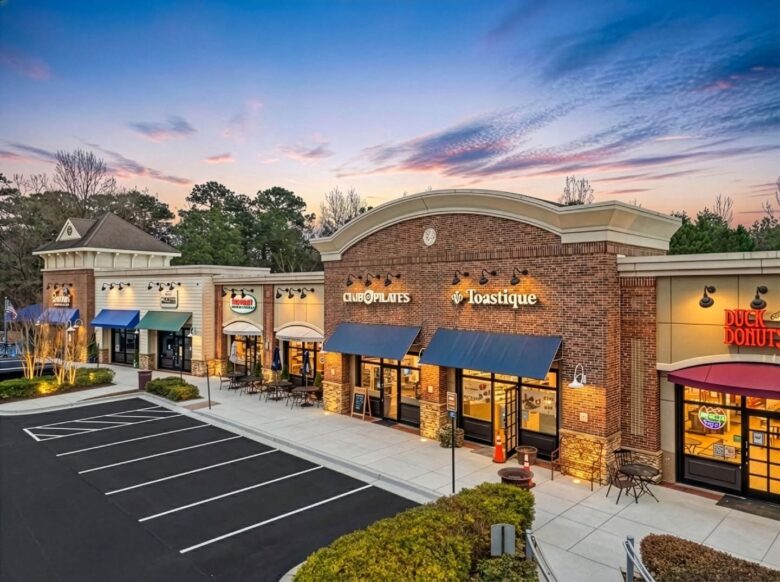 A small shopping plaza at dusk featuring several businesses with illuminated signs, including Club Pilates, Toastique, Duck Donuts, and others. The buildings have brick and light-colored facades with blue and green awnings. Outdoor seating with tables and chairs is visible on the sidewalk in front of some stores. The parking lot in front is empty, and trees are in the background under a partly cloudy sky with a sunset glow.