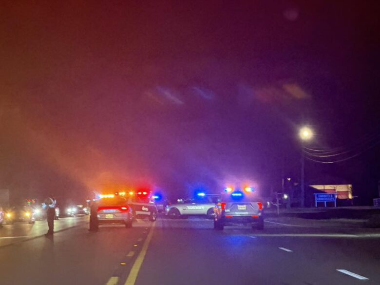 Multiple police vehicles with flashing red, blue, and yellow lights are blocking a road at night. A few people are standing near the vehicles, and other cars are visible in the background with their headlights on. The scene is illuminated by streetlights and emergency lights.
