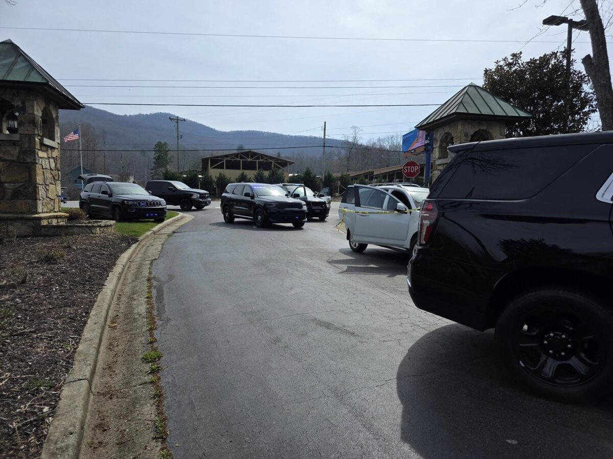 A group of black SUVs and a white car with its driver's side door open are parked on a paved road near stone gate structures with green metal roofs. The white car is surrounded by yellow caution tape. There is a stop sign and an American flag in the background, with hills and trees under a cloudy sky. Some of the black SUVs have blue lights on.