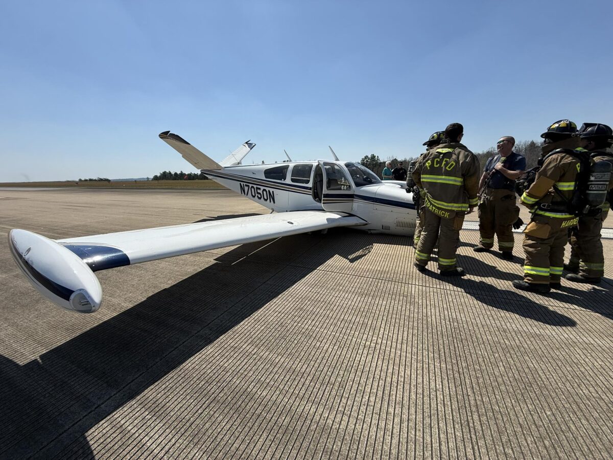 Small white airplane with blue stripes and the registration number N7050N parked on a concrete runway, with a group of firefighters in full gear standing nearby under a clear blue sky.