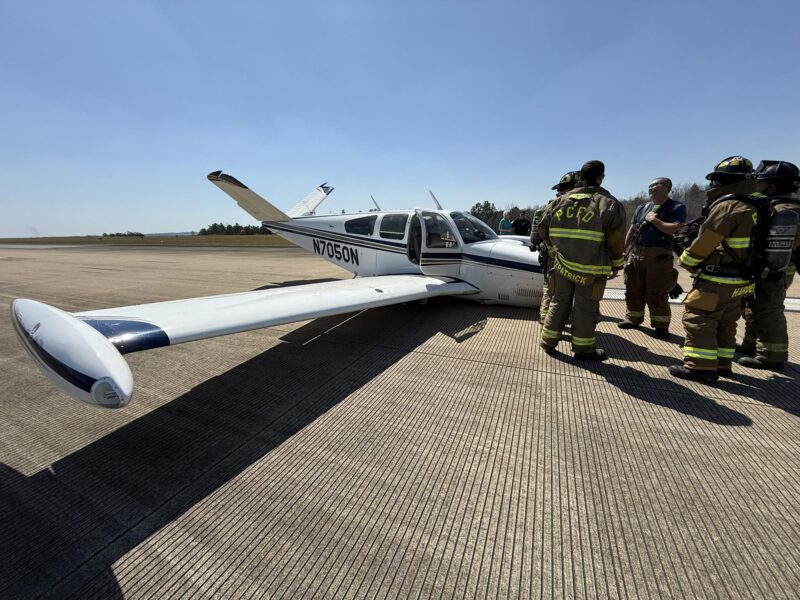 Small white airplane with blue stripes and the registration number N7050N parked on a concrete runway, with a group of firefighters in full gear standing nearby under a clear blue sky.