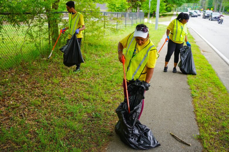 Three people wearing yellow safety vests and gloves are picking up litter along a roadside. Each person holds a large black trash bag and uses a grabber tool to collect trash from the grass and sidewalk. The background includes a chain-link fence, trees, and several vehicles on the road.