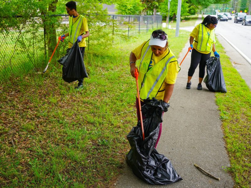 Three people wearing yellow safety vests and gloves are picking up litter along a roadside. Each person holds a large black trash bag and uses a grabber tool to collect trash from the grass and sidewalk. The background includes a chain-link fence, trees, and several vehicles on the road.